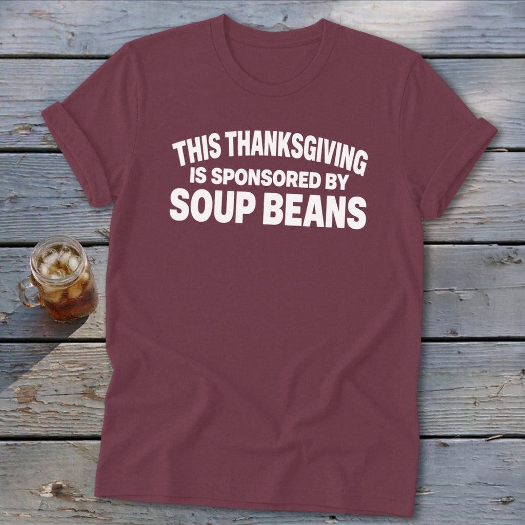 Maroon t-shirt with white text reading "This Thanksgiving is Sponsored by Soup Beans" laid on wooden planks.