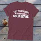 Maroon t-shirt with white text reading "This Thanksgiving is Sponsored by Soup Beans" laid on wooden planks.