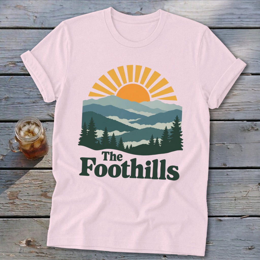 Pink t-shirt with a graphic of a sunrise over mountains and the text "The Foothills."