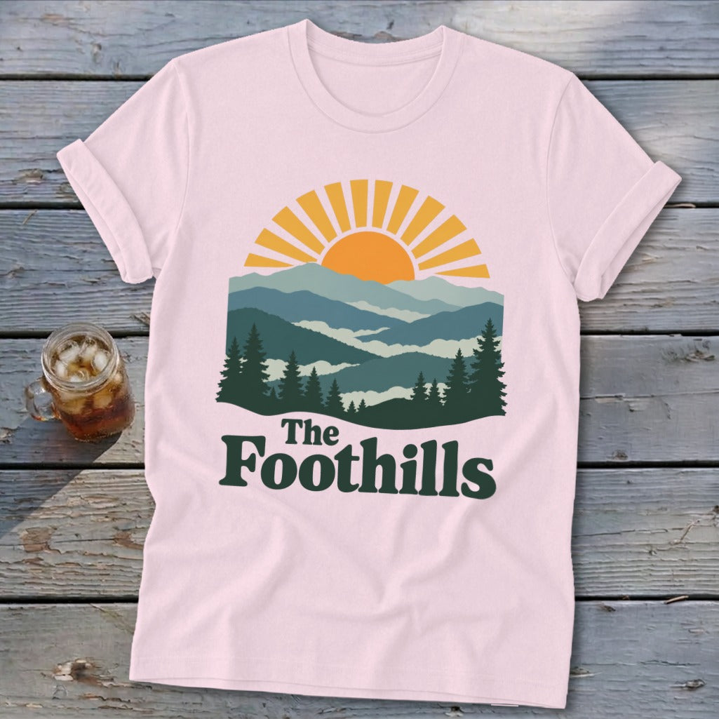 Pink t-shirt with a graphic of a sunrise over mountains and the text "The Foothills."