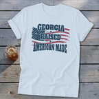 A white t-shirt featuring the text "GEORGIA RAISED AMERICAN MADE" over a distressed American flag graphic.