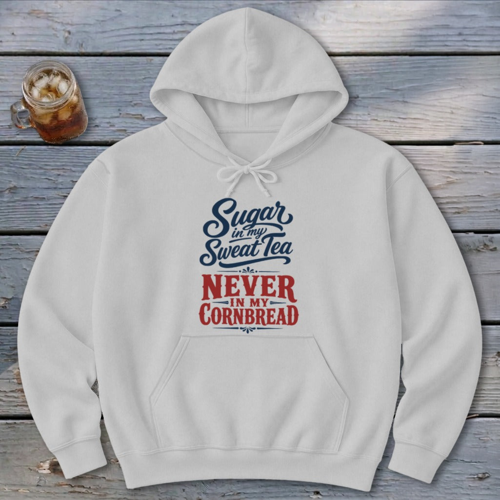 A light gray hooded sweatshirt featuring blue and red text reading Sugar in my Sweet Tea NEVER IN MY CORNBREAD rests on a wooden surface beside a glass of iced tea.