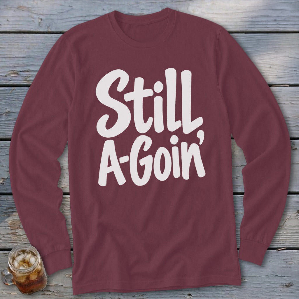 Maroon long-sleeve t-shirt with white text reading Still A-Goin' laid flat on a rustic wooden background.