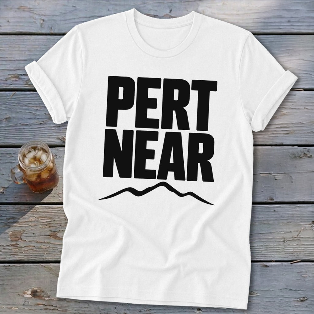 A white short-sleeved t-shirt with black block lettering reading PERT NEAR above a mountain range graphic, resting on a weathered wooden surface next to a glass of iced tea.