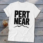 A white short-sleeved t-shirt with black block lettering reading PERT NEAR above a mountain range graphic, resting on a weathered wooden surface next to a glass of iced tea.