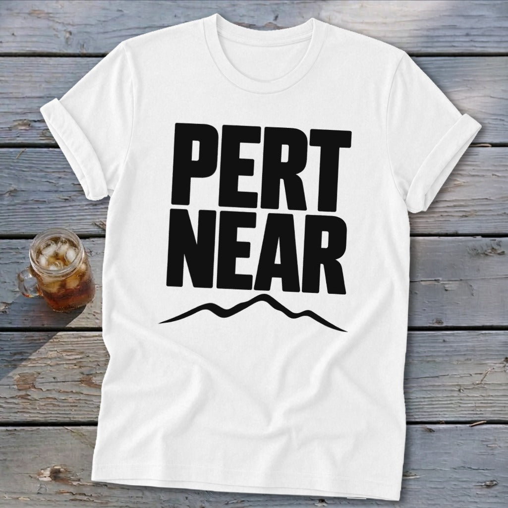 A white short-sleeved t-shirt with black block lettering reading PERT NEAR above a mountain range graphic, resting on a weathered wooden surface next to a glass of iced tea.