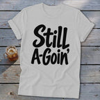 Heather gray crewneck t-shirt with rolled sleeves featuring the black text Still A-Goin' on a wooden background.