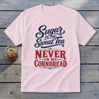 A light pink t-shirt with navy and red text that reads Sugar in my Sweat Tea, NEVER IN MY CORNBREAD laid flat on a rustic wooden table next to a glass of sweet tea.