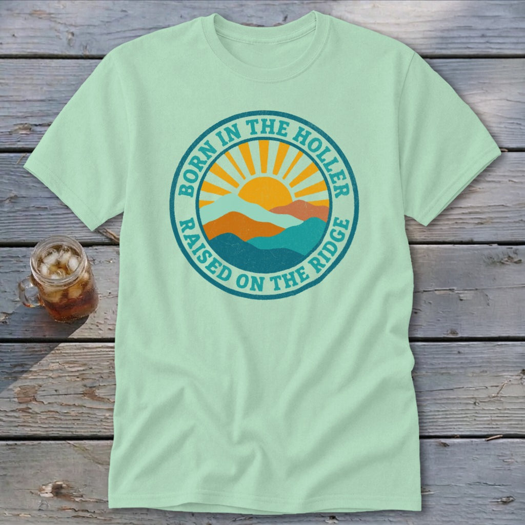 Light green short-sleeved t-shirt featuring a circular graphic with mountains, a rising sun, and the text Born in the Holler, Raised on the Ridge.