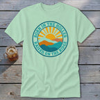 Light green short-sleeved t-shirt featuring a circular graphic with mountains, a rising sun, and the text Born in the Holler, Raised on the Ridge.