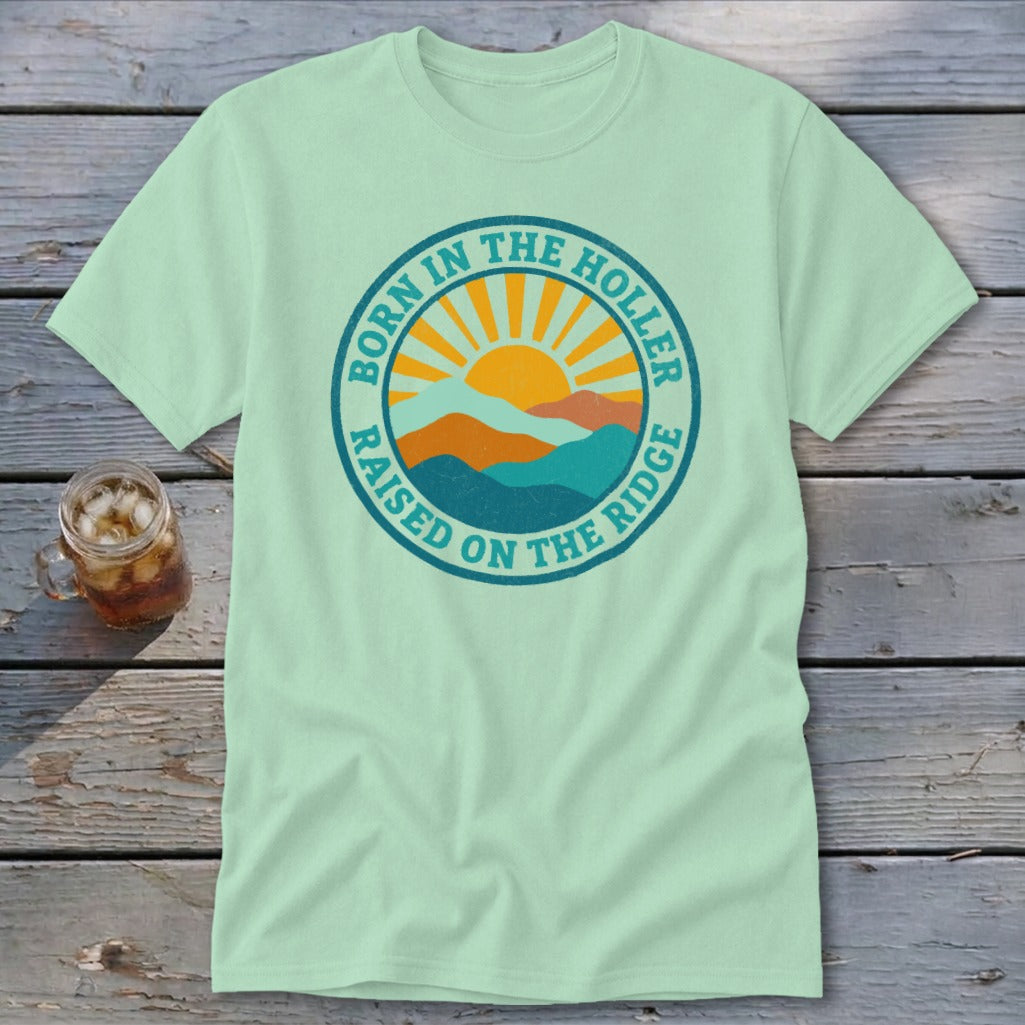 Light green short-sleeved t-shirt featuring a circular graphic with mountains, a rising sun, and the text Born in the Holler, Raised on the Ridge.