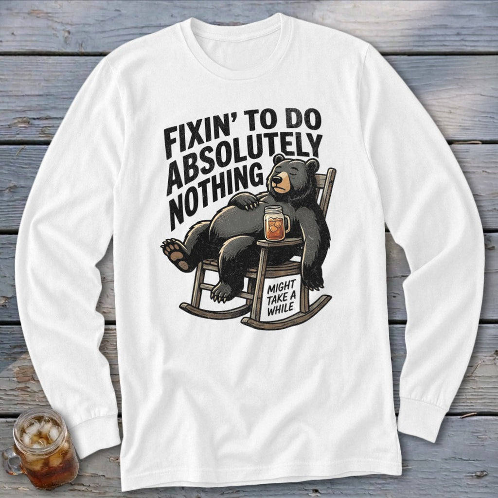 A white long-sleeved t-shirt with a graphic of a bear sleeping in a rocking chair, holding a drink, with the text "FIXIN' TO DO ABSOLUTELY NOTHING, MIGHT TAKE A WHILE."