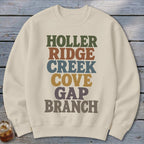 A cream-colored crewneck sweatshirt printed with stacked retro text reading HOLLER, RIDGE, CREEK, COVE, GAP, and BRANCH.