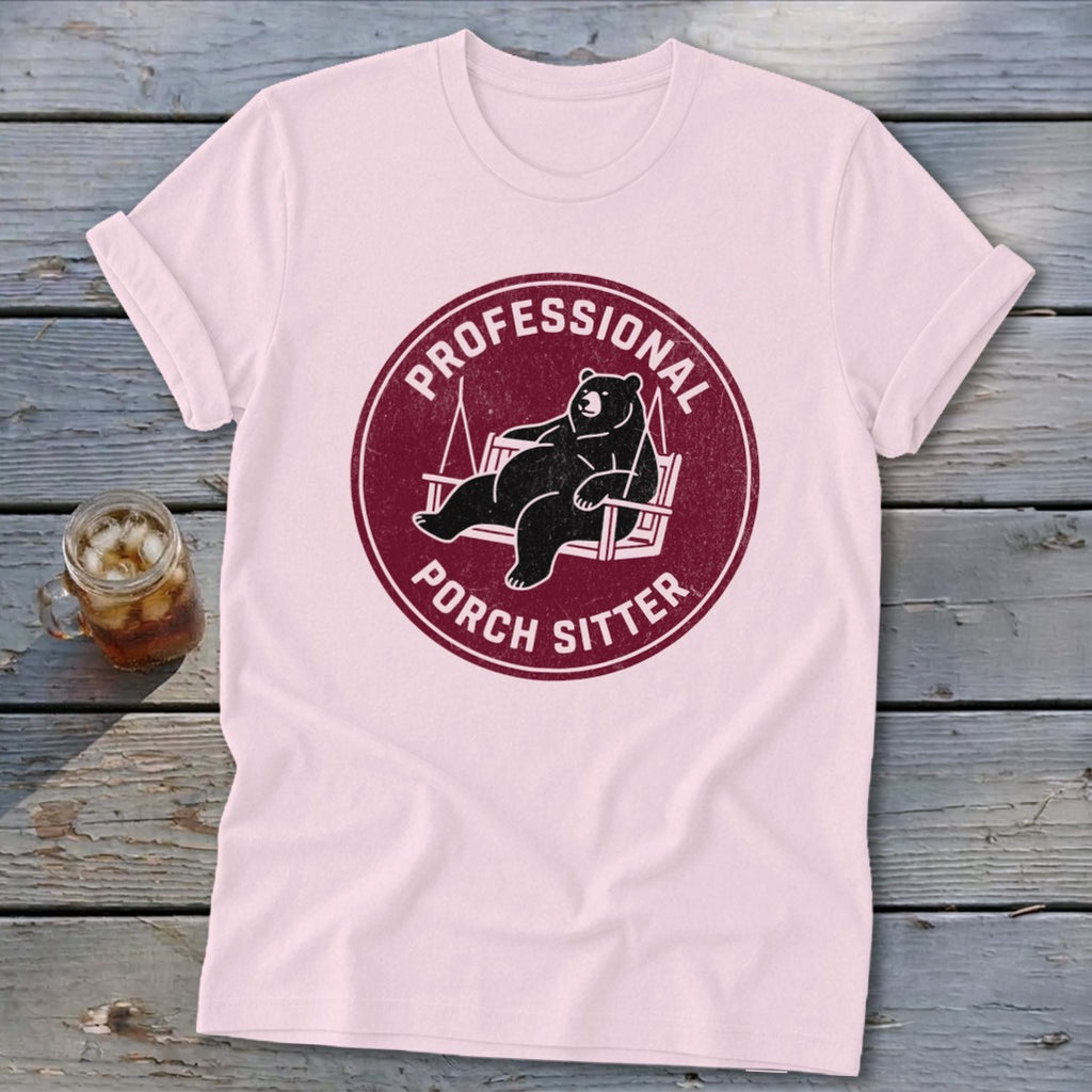 A light pink t-shirt with a maroon circular graphic featuring a bear relaxing on a swing, surrounded by the text PROFESSIONAL PORCH SITTER.