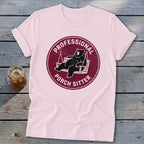 A light pink t-shirt with a maroon circular graphic featuring a bear relaxing on a swing, surrounded by the text PROFESSIONAL PORCH SITTER.