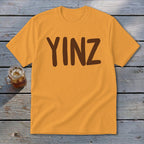 Orange t-shirt with brown "YINZ" text, set on wooden planks.
