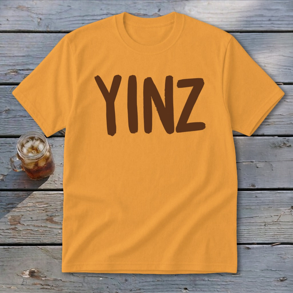 Orange t-shirt with brown "YINZ" text, set on wooden planks.