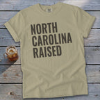 Tan t-shirt with "NORTH CAROLINA RAISED" printed in dark gray text.