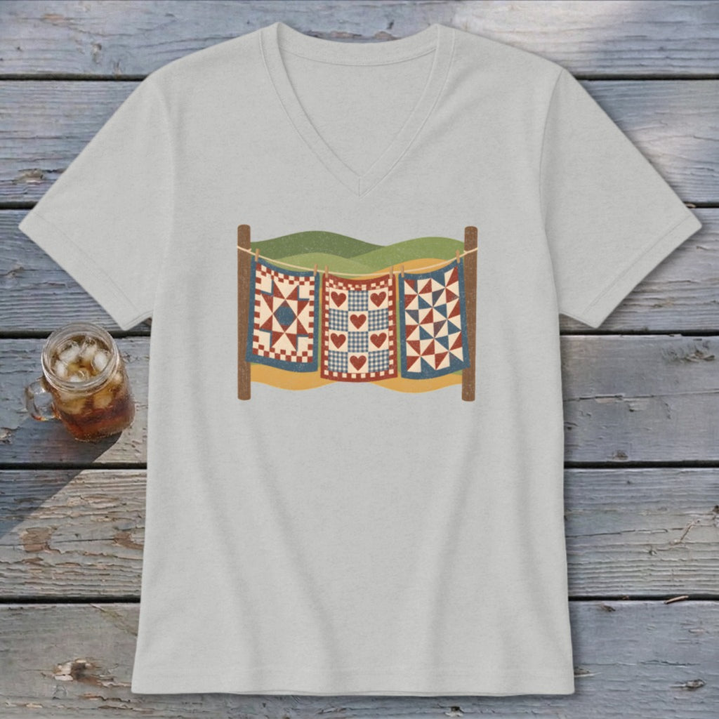A light gray V-neck t-shirt featuring a graphic of three patchwork quilts hanging on a clothesline against a landscape backdrop.