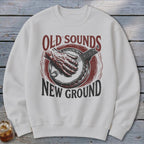 A white crewneck sweatshirt features a distressed red and black graphic of a hand playing a banjo, accompanied by the text OLD SOUNDS NEW GROUND.