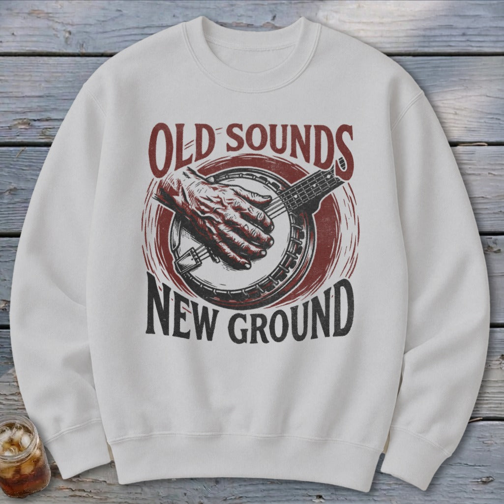 A white crewneck sweatshirt features a distressed red and black graphic of a hand playing a banjo, accompanied by the text OLD SOUNDS NEW GROUND.