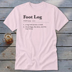 Light pink short-sleeve t-shirt with black dictionary definition text printed on the chest, laid flat on a rustic wooden table next to a glass of iced tea.