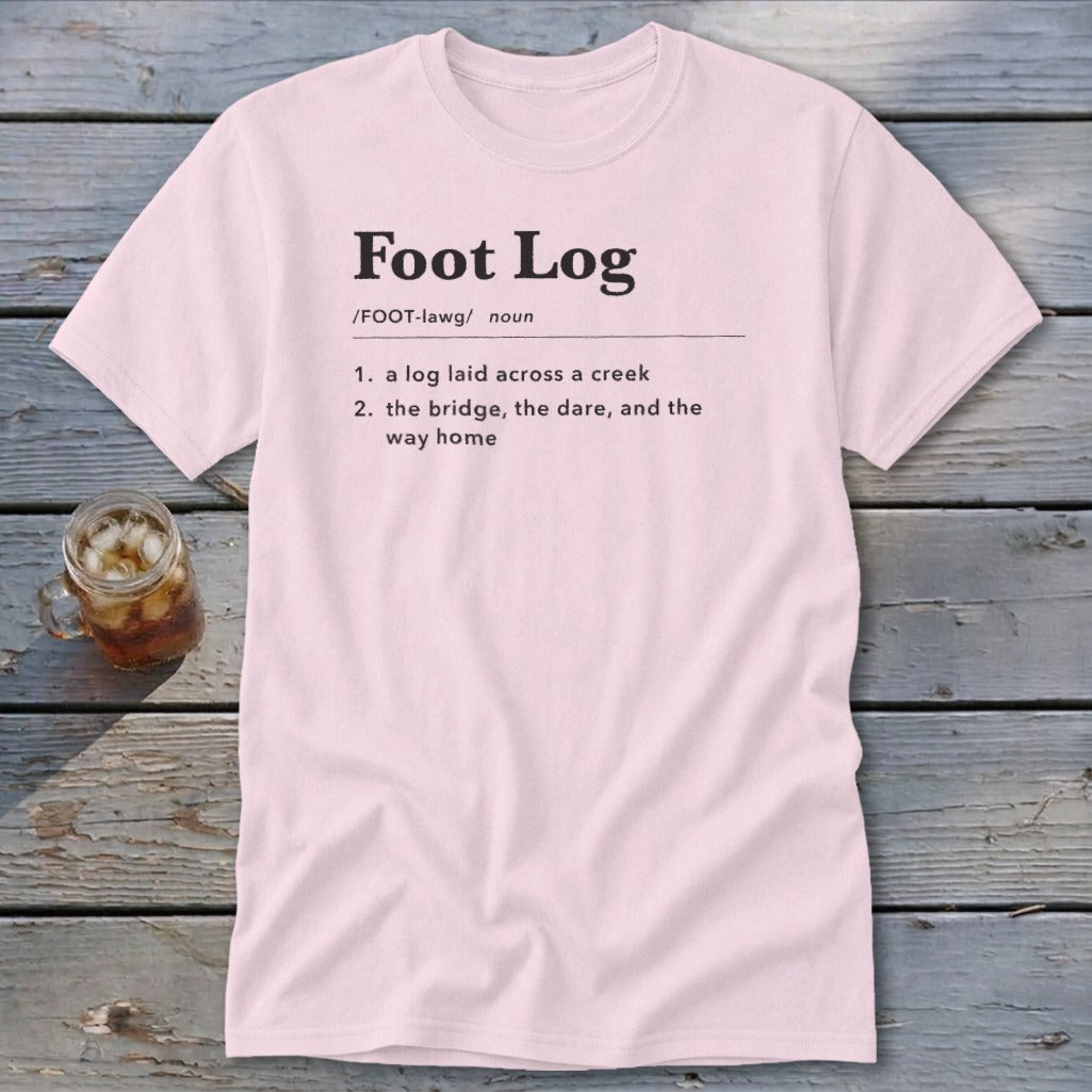 Light pink short-sleeve t-shirt with black dictionary definition text printed on the chest, laid flat on a rustic wooden table next to a glass of iced tea.