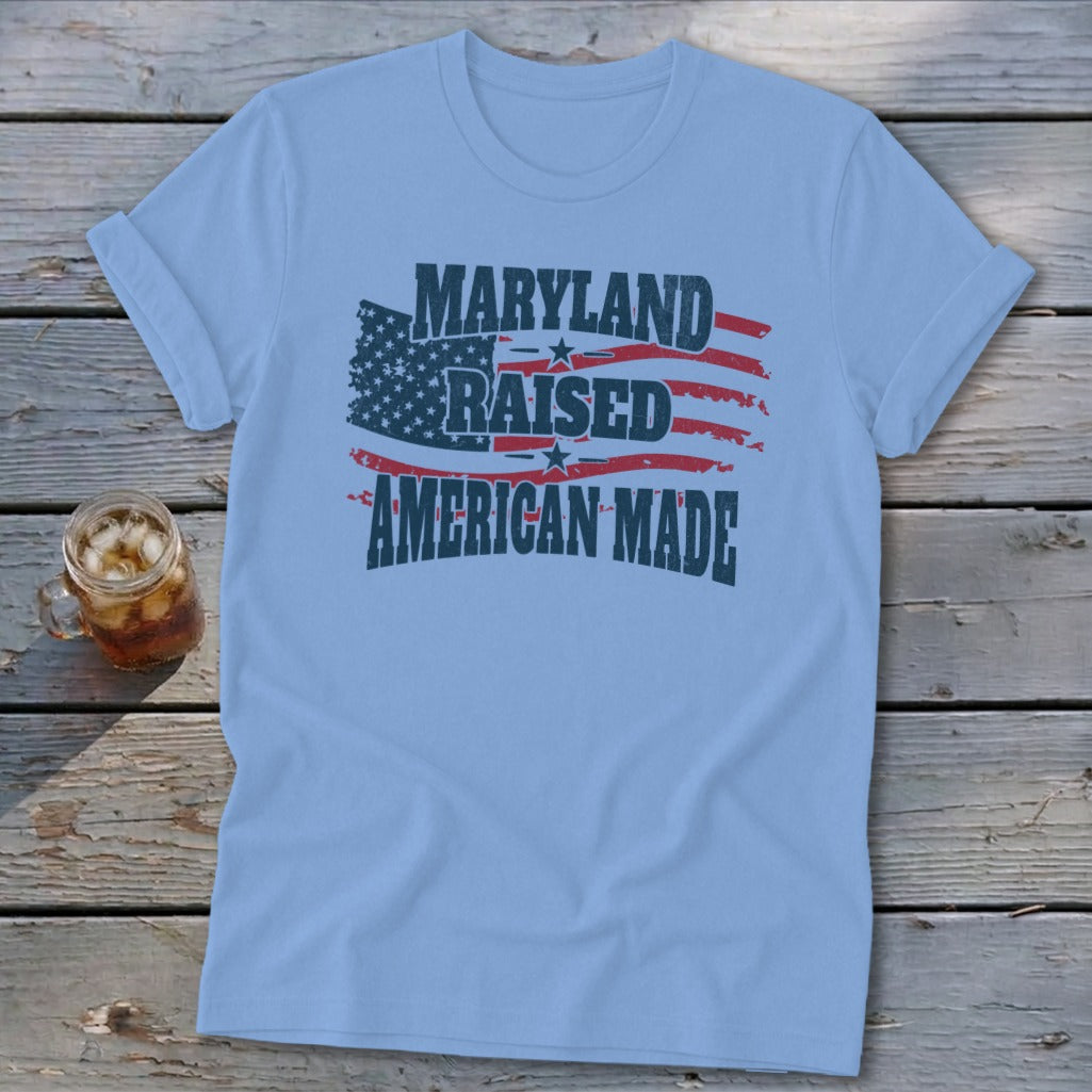 Light blue t-shirt featuring distressed text reading "MARYLAND RAISED AMERICAN MADE" and a stylized American flag graphic.
