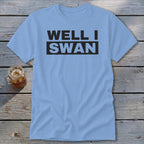 Light blue t-shirt featuring the black text WELL I SWAN laid on a wooden surface.