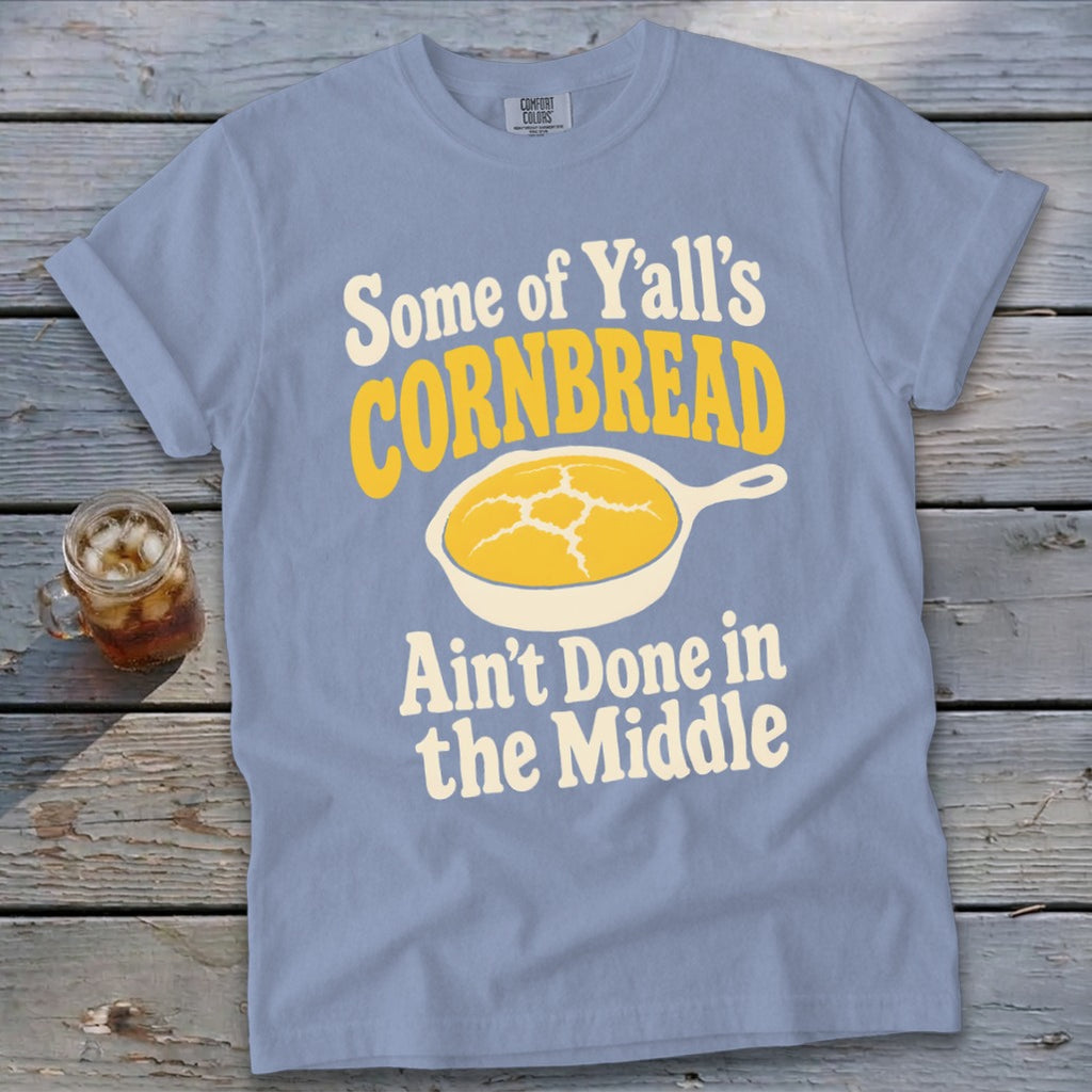 A light blue t-shirt with text that reads Some of Y'all's Cornbread Ain't Done in the Middle and a graphic of a skillet with cornbread.
