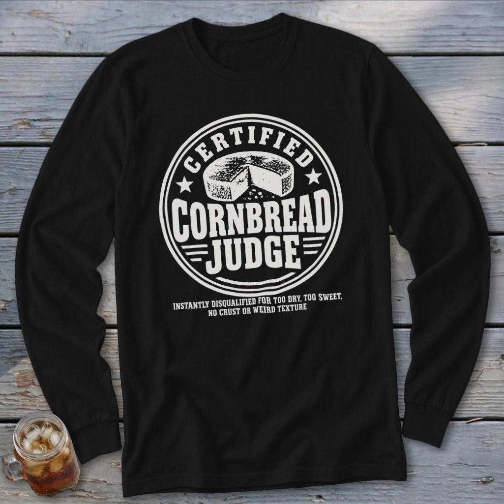 A black long-sleeve t-shirt with a white graphic featuring a round seal, cornbread illustration, and text reading Certified Cornbread Judge.