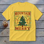 A yellow t-shirt features a vintage-style Christmas tree graphic with the text Mountain Merry on a wooden surface.