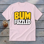 Light pink crewneck t-shirt featuring the text BUM FUZZLED in yellow and white on a weathered wooden surface.