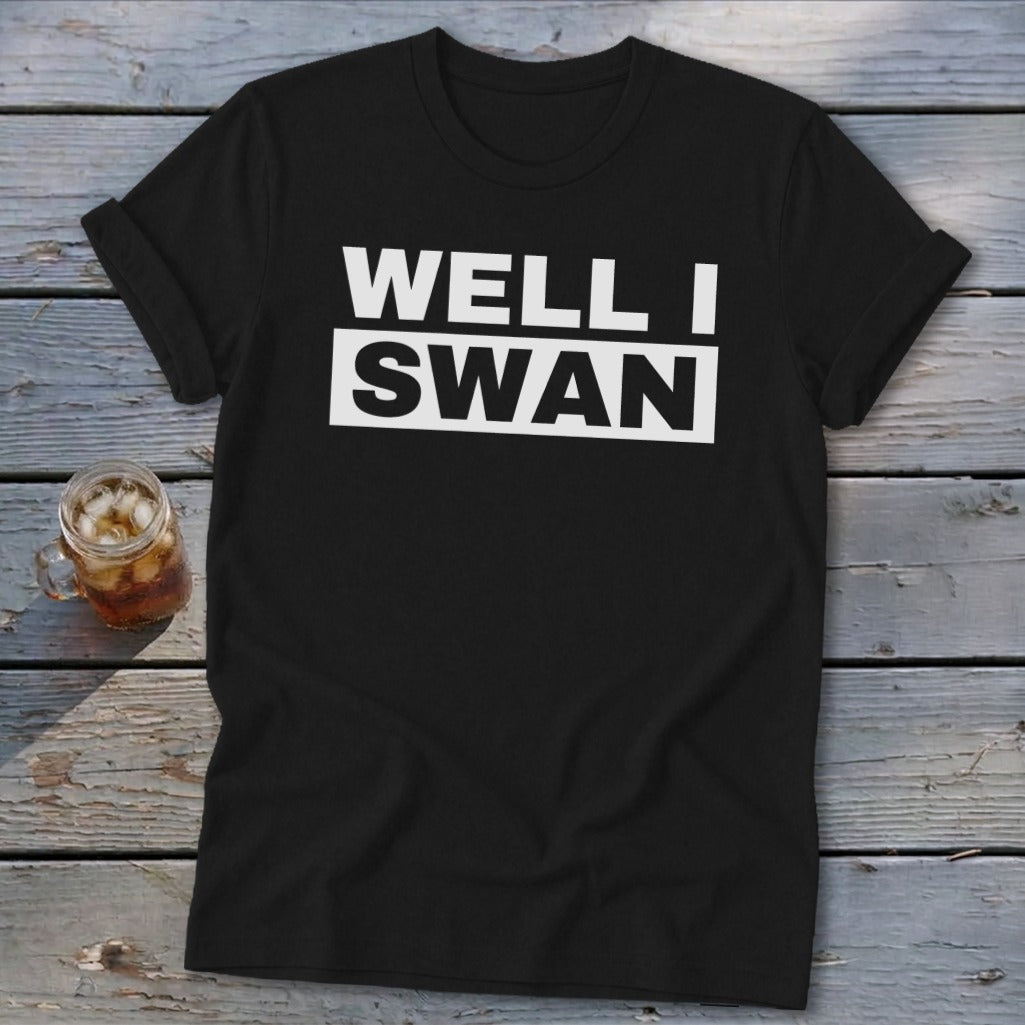 A black short-sleeved t-shirt with white block text reading Well I Swan across the chest rests on a weathered wooden surface next to a glass of iced tea.