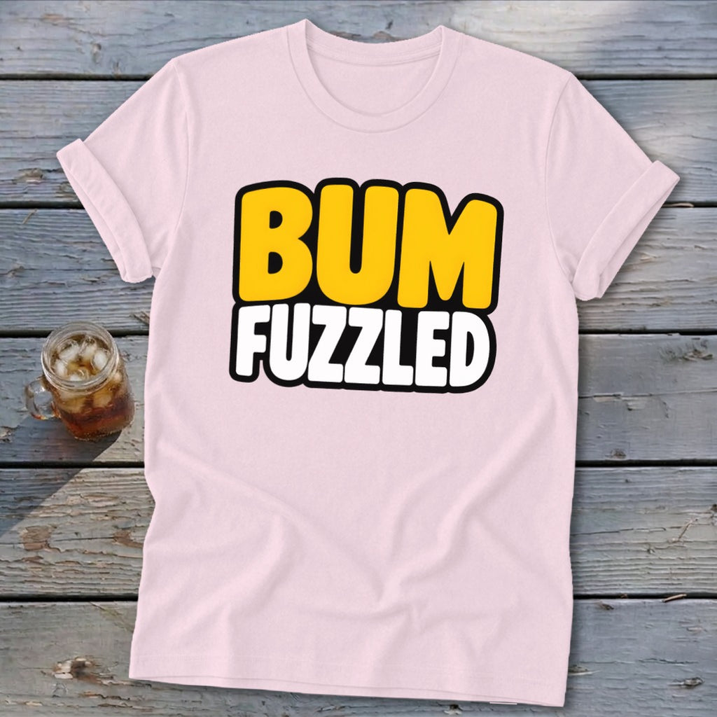 Light pink short-sleeve t-shirt with the text BUM FUZZLED on the chest, lying on a wooden surface next to a glass of iced tea.