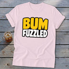 Light pink short-sleeve t-shirt with the text BUM FUZZLED on the chest, lying on a wooden surface next to a glass of iced tea.