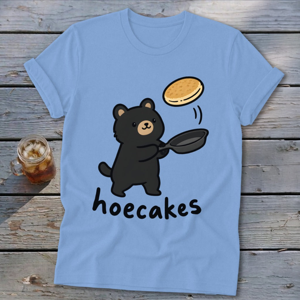 Light blue t-shirt featuring a cartoon black bear flipping a pancake in a pan, with the text hoecakes underneath.