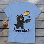 Light blue t-shirt featuring a cartoon black bear flipping a pancake in a pan, with the text hoecakes underneath.