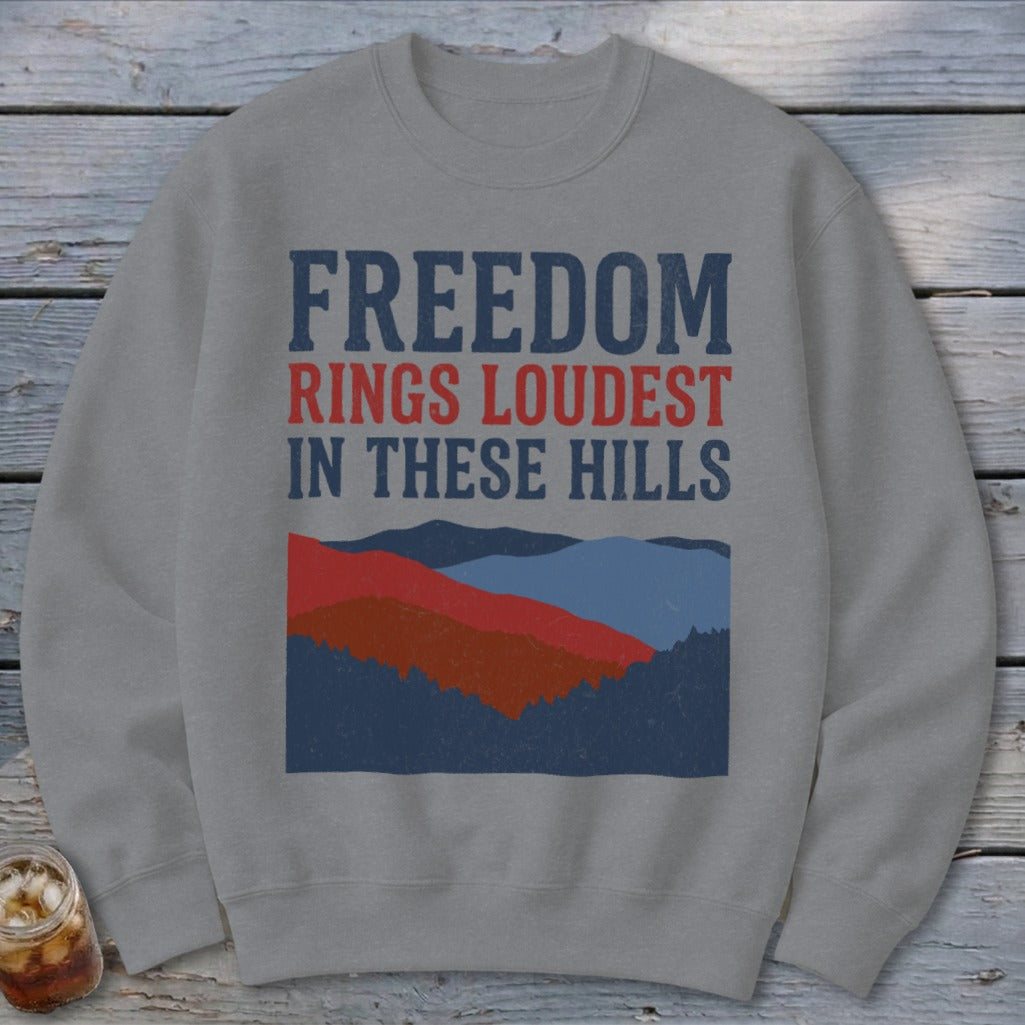 A gray crewneck sweatshirt printed with the text FREEDOM RINGS LOUDEST IN THESE HILLS above a mountain illustration.