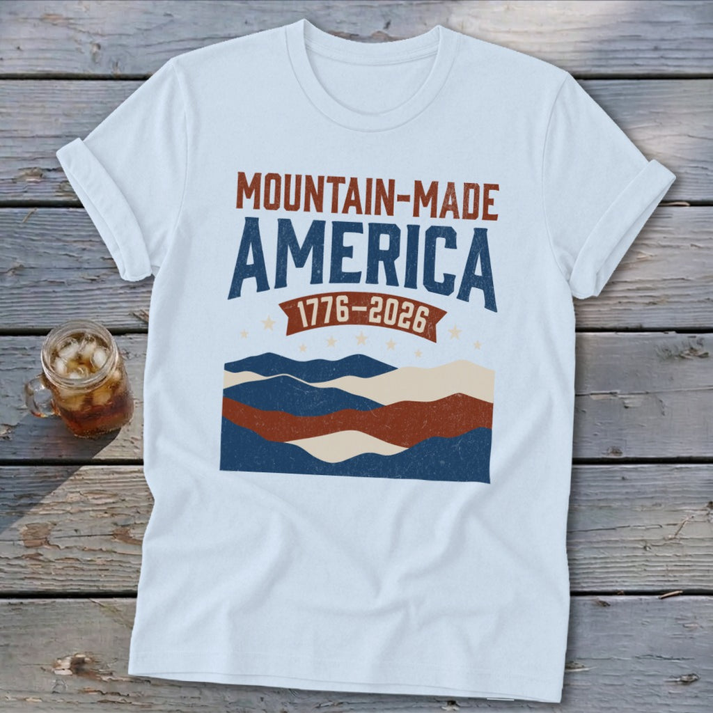 A white t-shirt featuring a graphic with the text "MOUNTAIN-MADE AMERICA 1776-2026" above stylized mountains and stars.