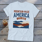 A white t-shirt featuring a graphic with the text "MOUNTAIN-MADE AMERICA 1776-2026" above stylized mountains and stars.