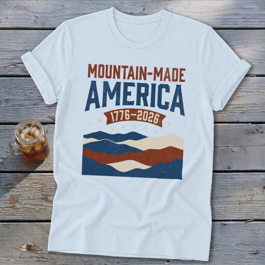 A white t-shirt featuring a graphic with the text "MOUNTAIN-MADE AMERICA 1776-2026" above stylized mountains and stars.