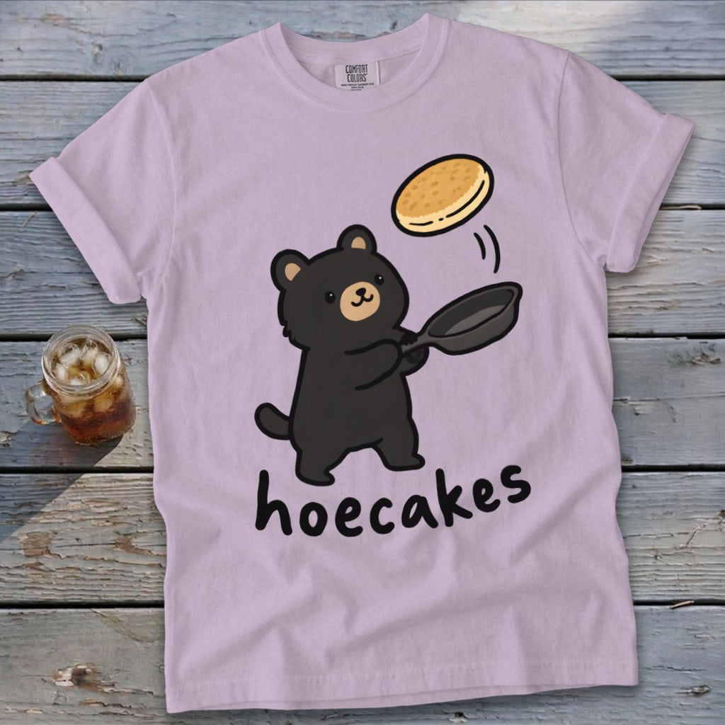 Light purple t-shirt featuring a cartoon bear flipping a pancake in a pan with the text hoecakes.