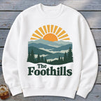 White sweatshirt with mountain landscape graphic and "The Foothills" text.
