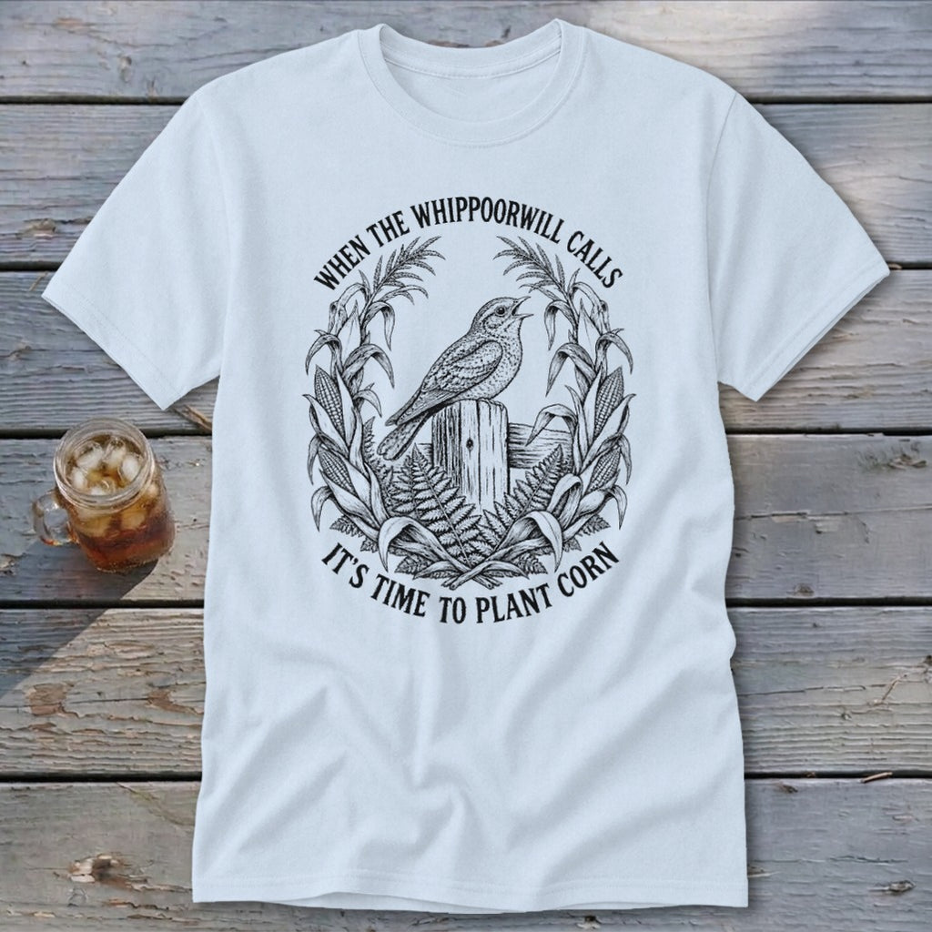 White crewneck t-shirt featuring a black graphic of a bird perched on a wooden post, surrounded by corn stalks and the text "When the whippoorwill calls, it's time to plant corn."