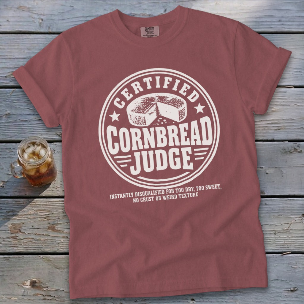 A dusty rose crewneck t-shirt with white graphic text reading Certified Cornbread Judge and an illustration of a cornbread wheel.