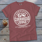 A dusty rose crewneck t-shirt with white graphic text reading Certified Cornbread Judge and an illustration of a cornbread wheel.