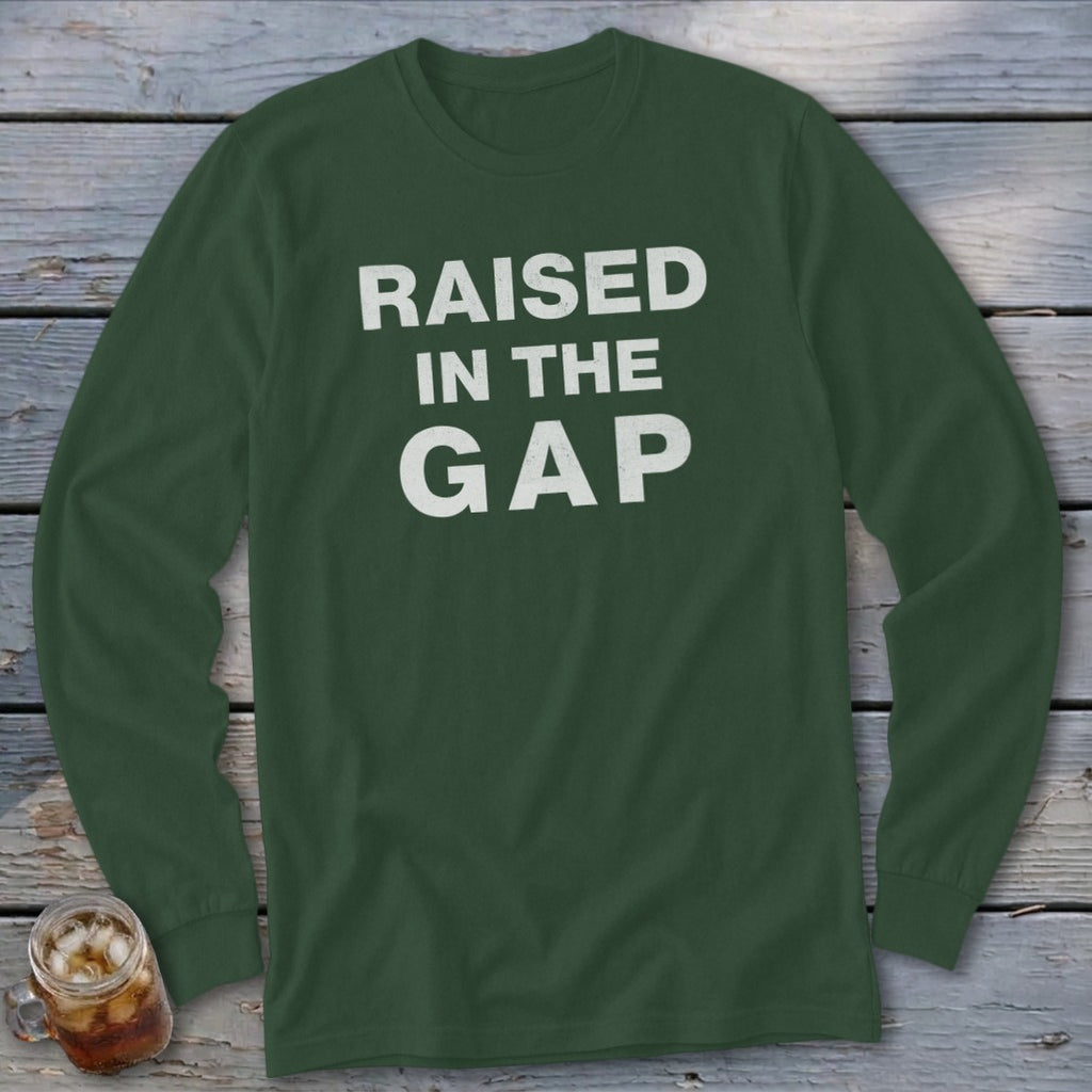 Dark green long-sleeve t-shirt with white text reading "RAISED IN THE GAP" centered on the chest, lying on a wooden surface.