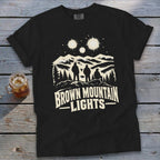 Black t-shirt featuring a graphic of three figures viewing glowing mountain lights above a forest landscape.