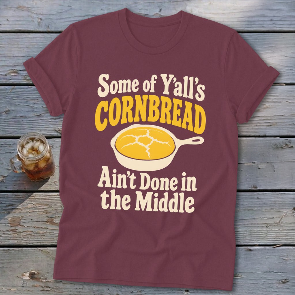 Maroon t-shirt featuring text and a skillet graphic with cornbread, laid flat on a wooden surface.